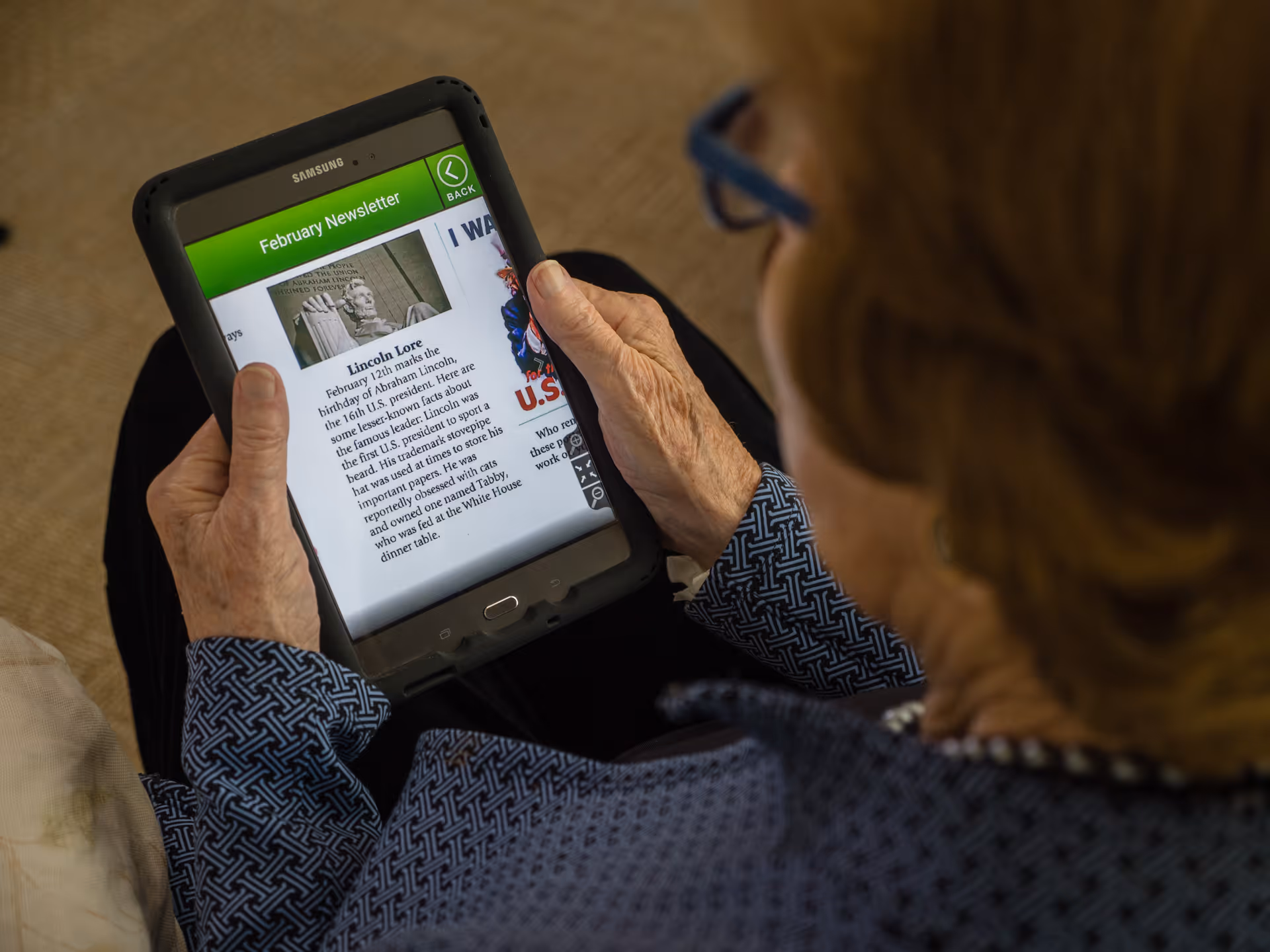 An elderly person wearing glasses and a patterned blue shirt is holding a Samsung tablet displaying a February newsletter with an article titled 'Lincoln Lore' and an image of a Lincoln statue.