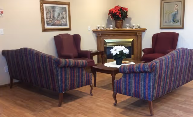 A cozy living room area with two striped sofas facing each other and two maroon armchairs positioned near a wooden fireplace mantel. The mantel is decorated with a red poinsettia plant and several small candles. A coffee table with a white poinsettia plant on a doily is centered between the sofas. The walls are adorned with framed artwork, and the floor is wooden.