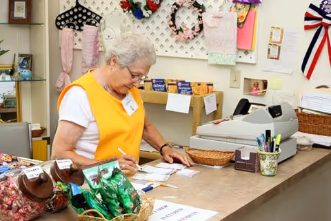 An elderly woman wearing a yellow vest and white shirt is writing on papers at a counter in a room decorated with various crafts and items. The counter has baskets with snacks and office supplies, and a cash register is visible in the background.