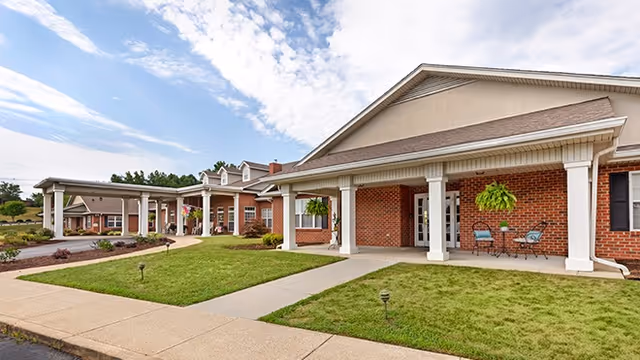 Brick single-story senior living facility entrance with white columns, covered walkways, hanging plants, and a paved path across a lawn.