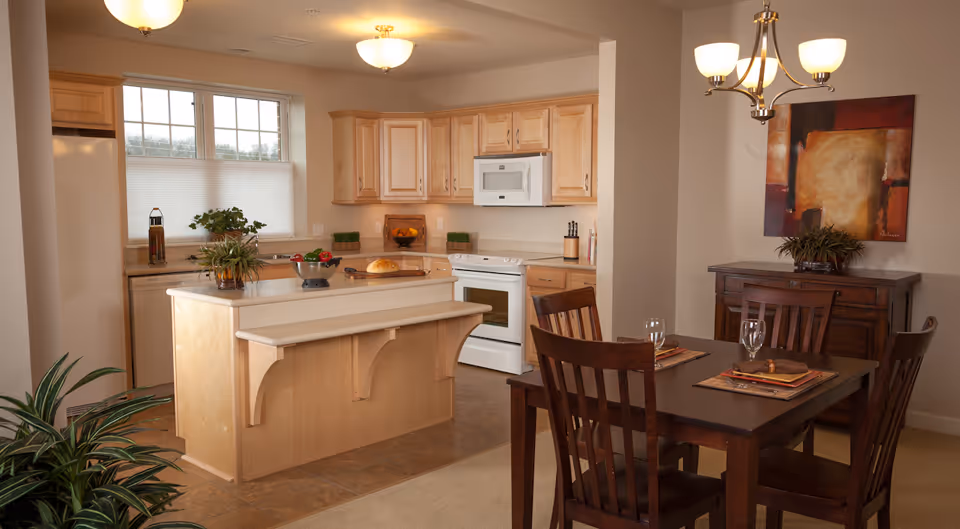 A kitchen and dining area in a senior living facility. The kitchen features light wood cabinets, a white stove, microwave, and refrigerator. There is a kitchen island with a small counter extension and plants on top. The dining area has a dark wood table set with four chairs, placemats, and glasses. A sideboard with a plant and abstract artwork hangs on the wall above it. The space is warmly lit with ceiling lights and a chandelier.