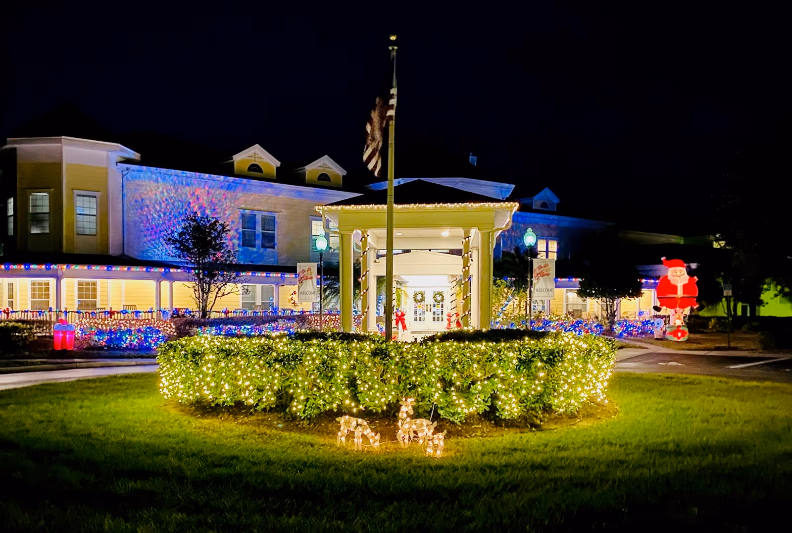 Night view of a decorated senior living entrance with holiday lights, wreaths, and an inflatable Santa on the lawn.