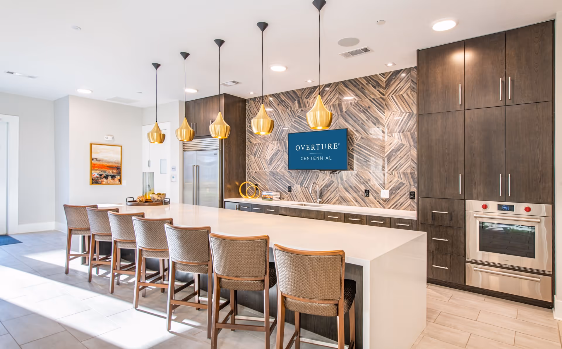 Modern kitchen area with a long white island countertop surrounded by six cushioned bar stools. Above the island hang five gold pendant lights. The back wall features a geometric patterned tile backsplash, dark wood cabinets, a built-in stainless steel oven, and a large stainless steel refrigerator. A TV mounted on the wall displays the text 'OVERTURE CENTENNIAL'.