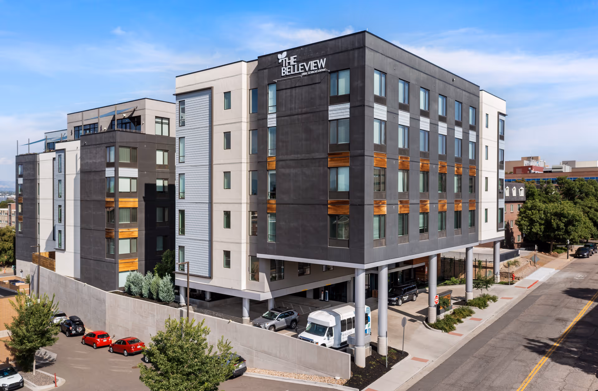 Exterior view of The Belleview Senior Living building, a modern multi-story structure with a combination of dark and light facade panels, large windows, and wooden accents. The building is elevated on pillars with parking spaces underneath and is situated along a street with trees and parked cars.