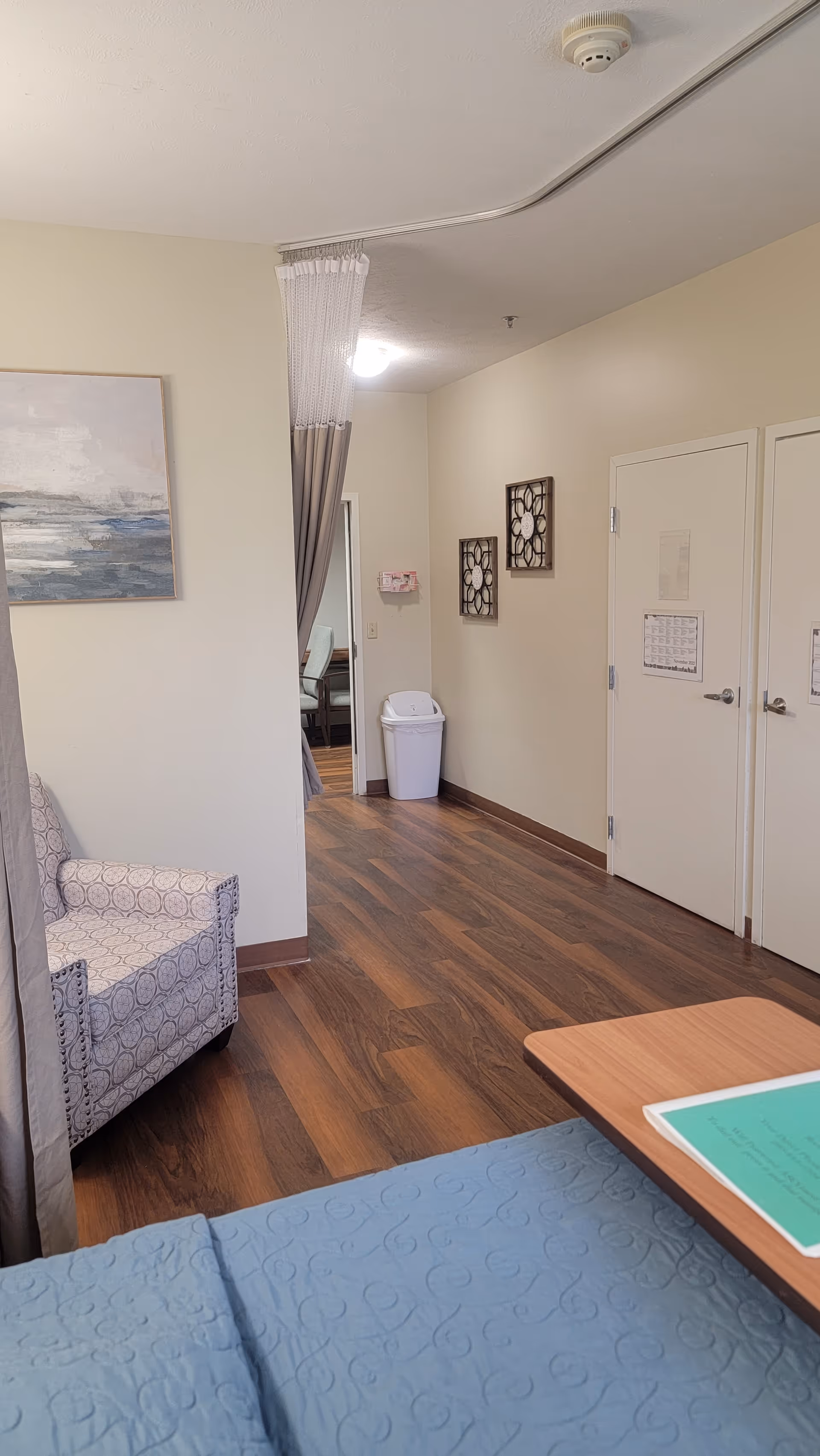 Interior view of a senior living facility room at Coventry Meadows showing a patterned armchair, a bed with a blue quilt, a wooden table with a green folder, wooden flooring, two closed white doors with notices, wall decorations, a trash bin, and a partially visible adjoining room with chairs.