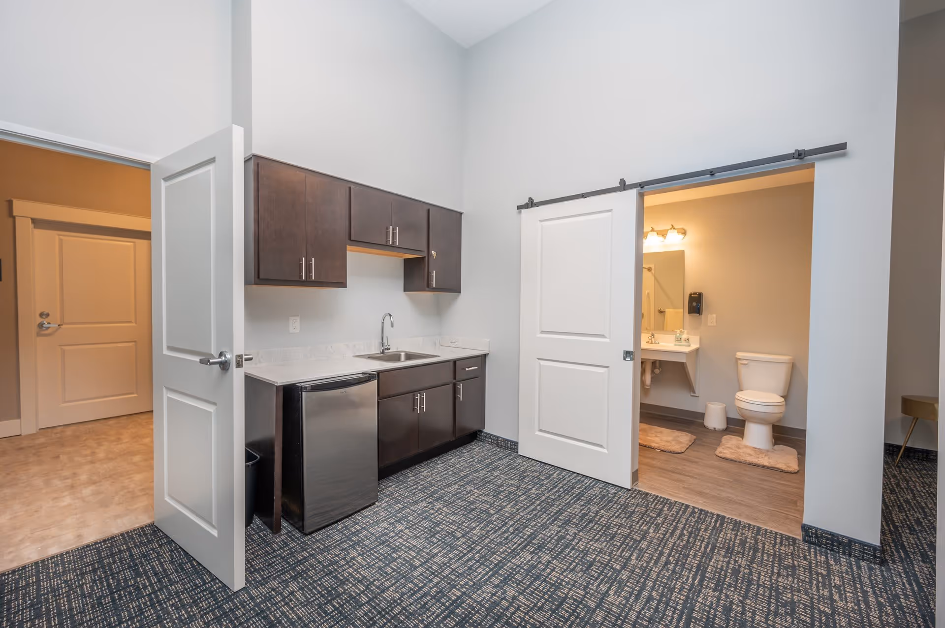 Interior view of a room with a small kitchenette featuring dark wood cabinets, a stainless steel mini fridge, and a sink. To the right, a sliding door is open, revealing a bathroom with a toilet, sink, mirror, and wall-mounted soap dispenser. The room has patterned carpet flooring and light gray walls.