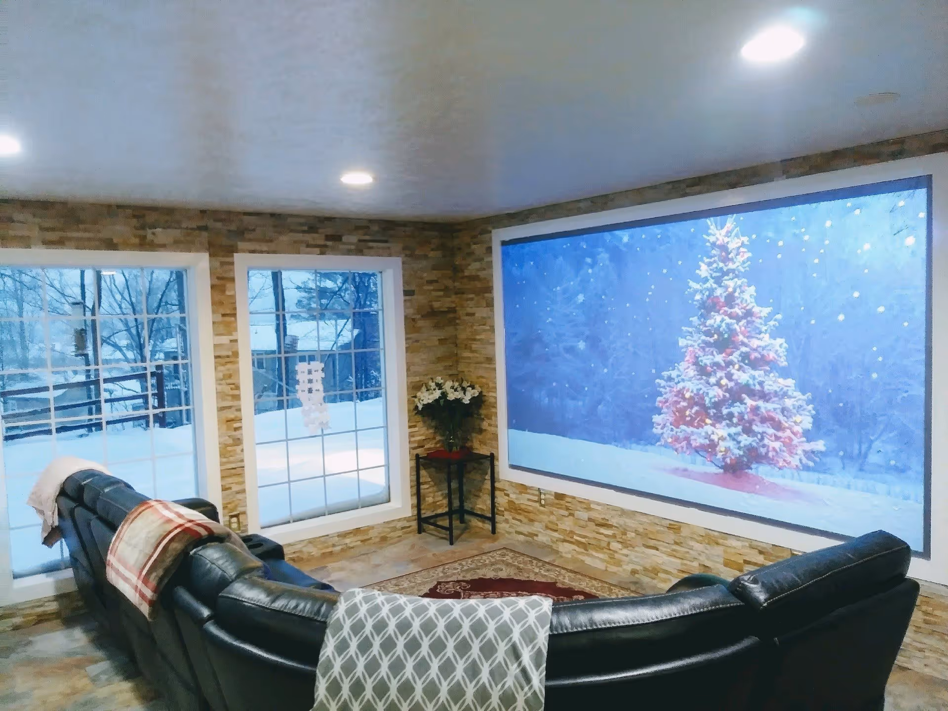 A cozy living room with a large black leather sectional sofa adorned with patterned blankets. The room has stone-textured walls and two large windows showing a snowy outdoor scene. A large screen on the wall displays a snowy Christmas tree with colorful lights. A small table with a flower arrangement is placed in the corner.