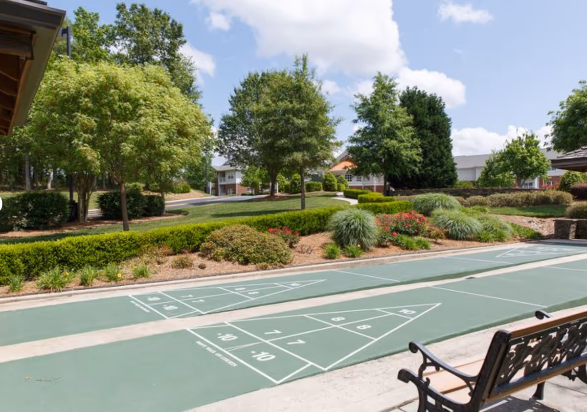 Outdoor shuffleboard courts with a bench, landscaped shrubs and trees, and buildings in the background.