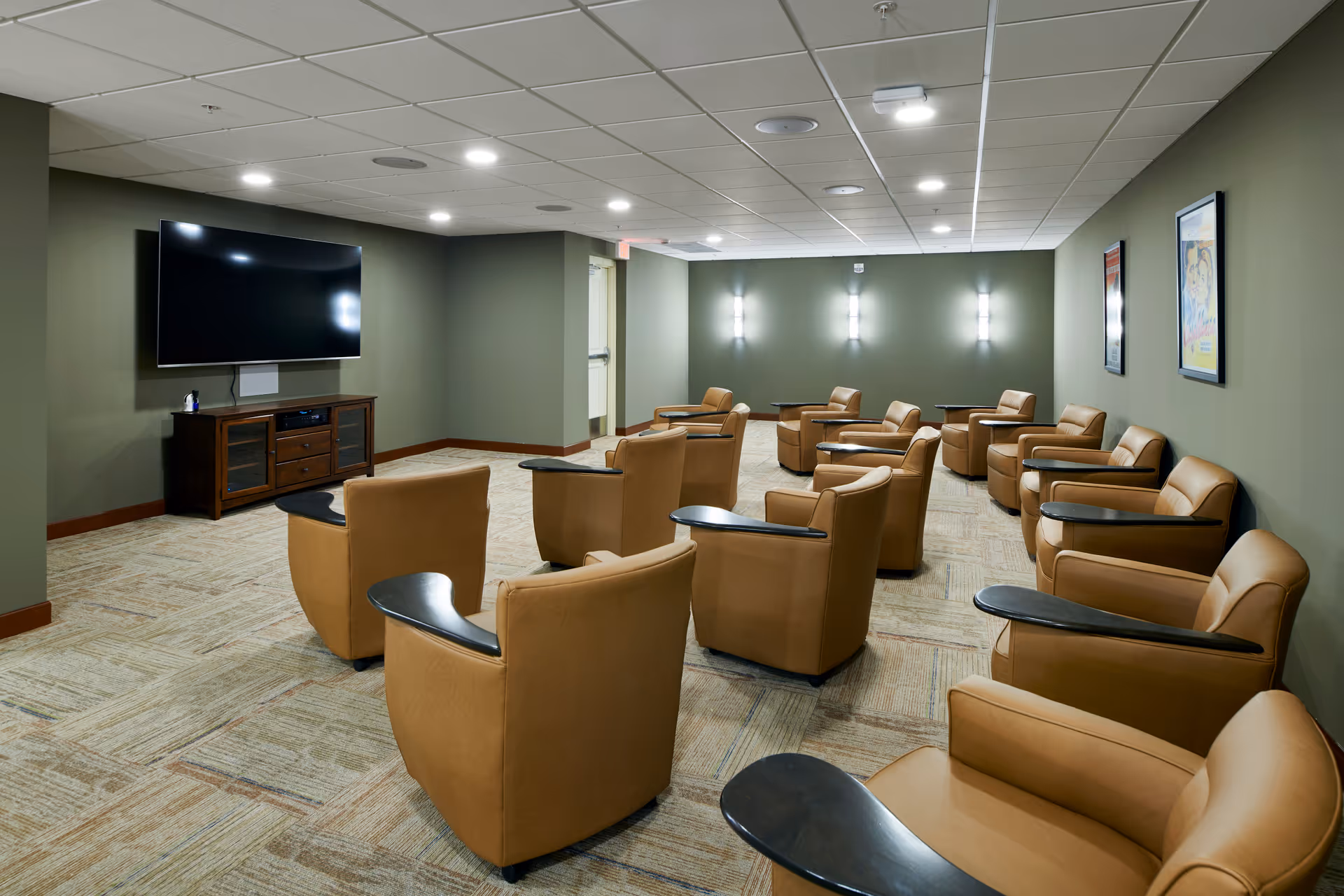 A media-style lounge with rows of tan armchairs facing a wall-mounted TV in a green-walled room.