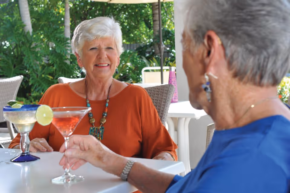 Two elderly women sitting at an outdoor table enjoying drinks, one woman in an orange top smiling and the other in a blue top holding a cocktail glass with a pink drink.