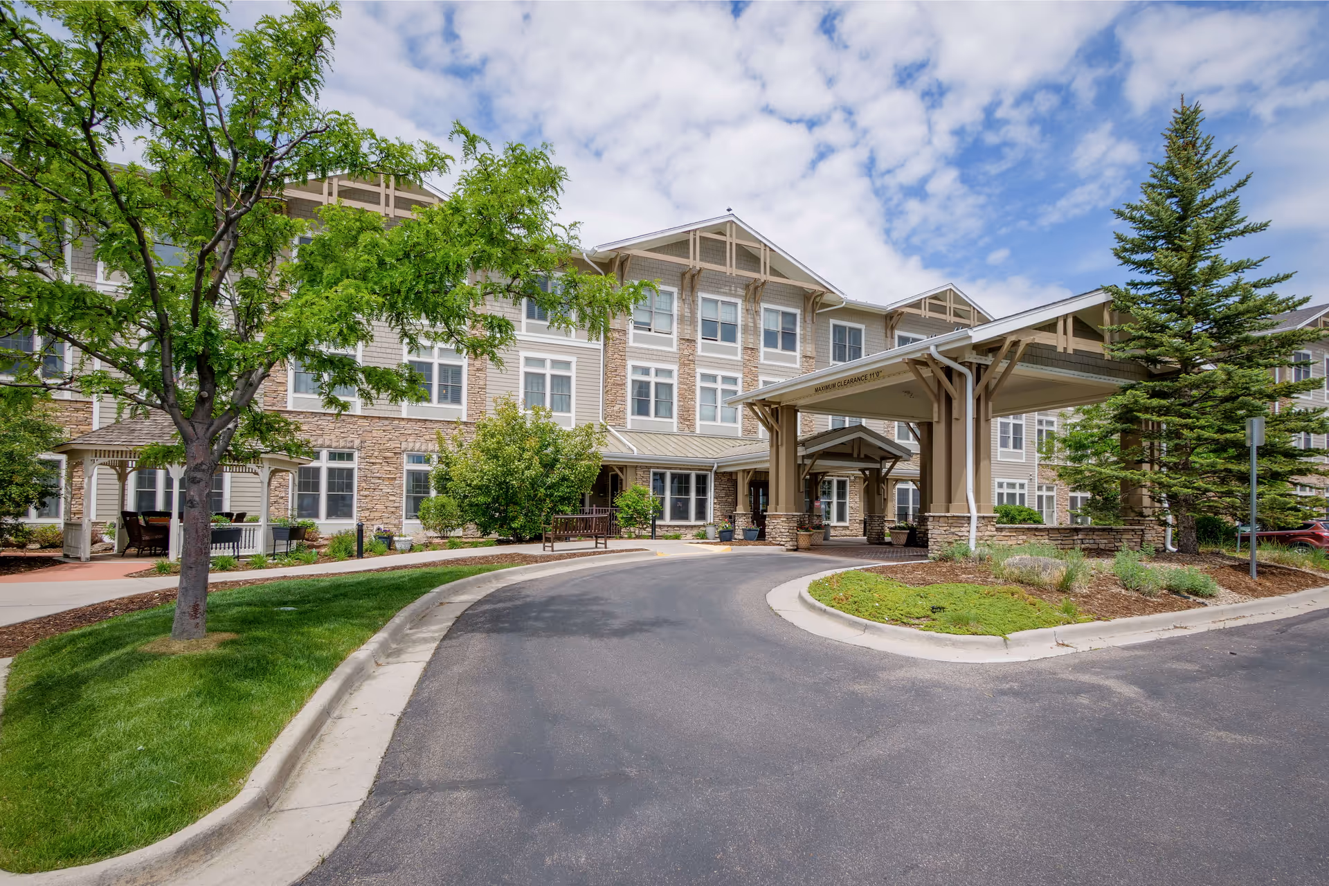 Exterior view of Sunrise at FlatIrons senior living facility showing a three-story building with a covered entrance driveway, landscaped greenery including trees and shrubs, and a partly cloudy sky.
