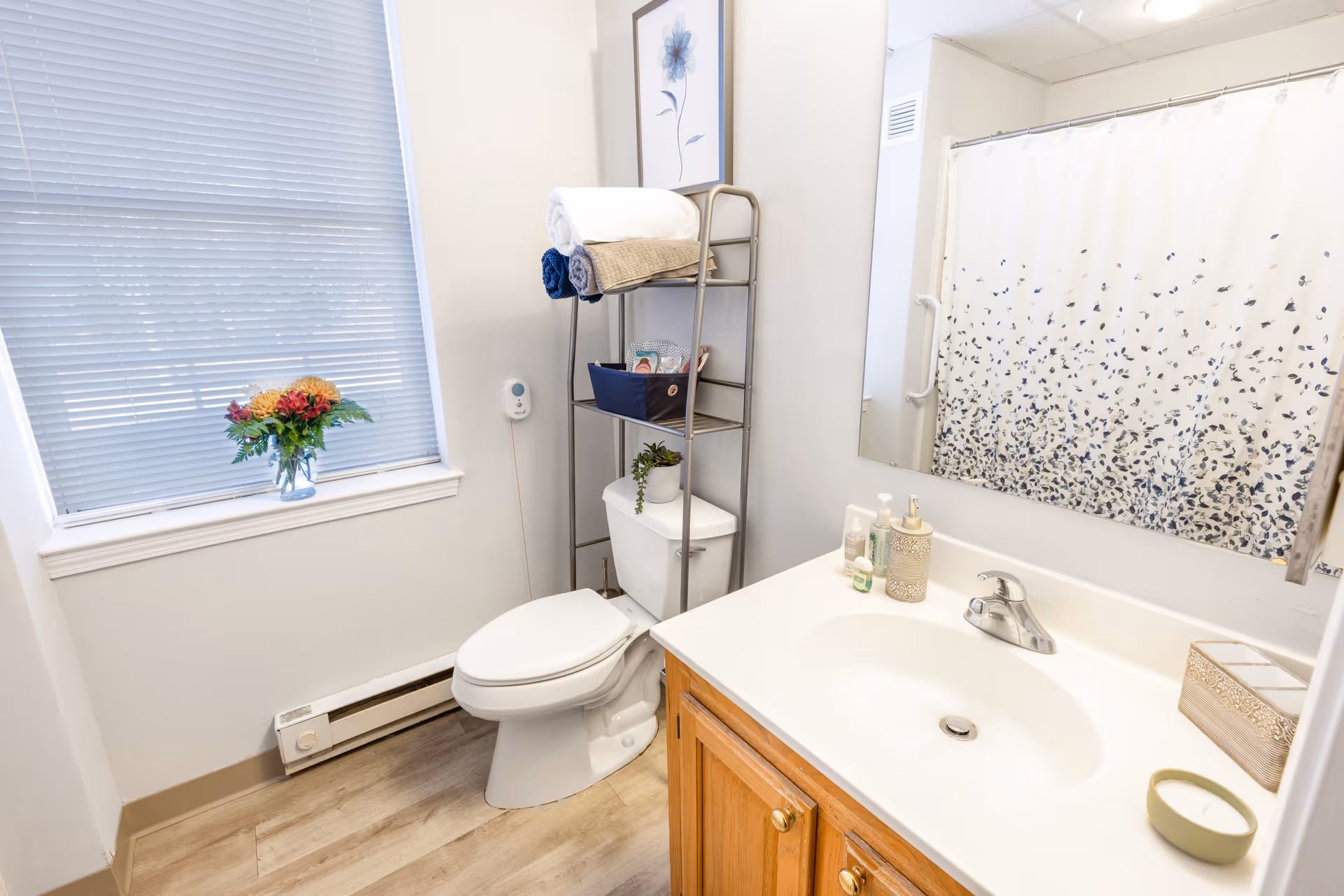 A clean bathroom with a white toilet, a metal shelving unit holding towels and toiletries, a wooden vanity with a white sink and faucet, a large mirror above the sink, a shower with a white curtain featuring a blue floral pattern, a window with closed blinds, and a small vase of flowers on the windowsill.