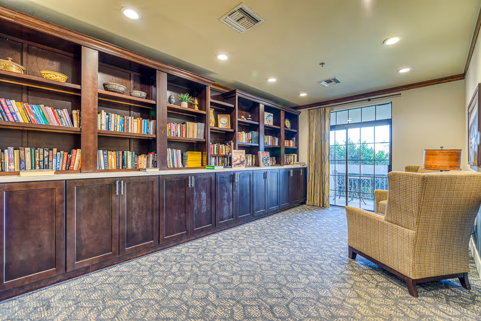 A cozy reading room with a long dark wooden bookshelf filled with books and decorative items. Two beige upholstered armchairs are positioned near a large window with glass doors leading to an outdoor balcony. The room has patterned carpet flooring and recessed ceiling lights.