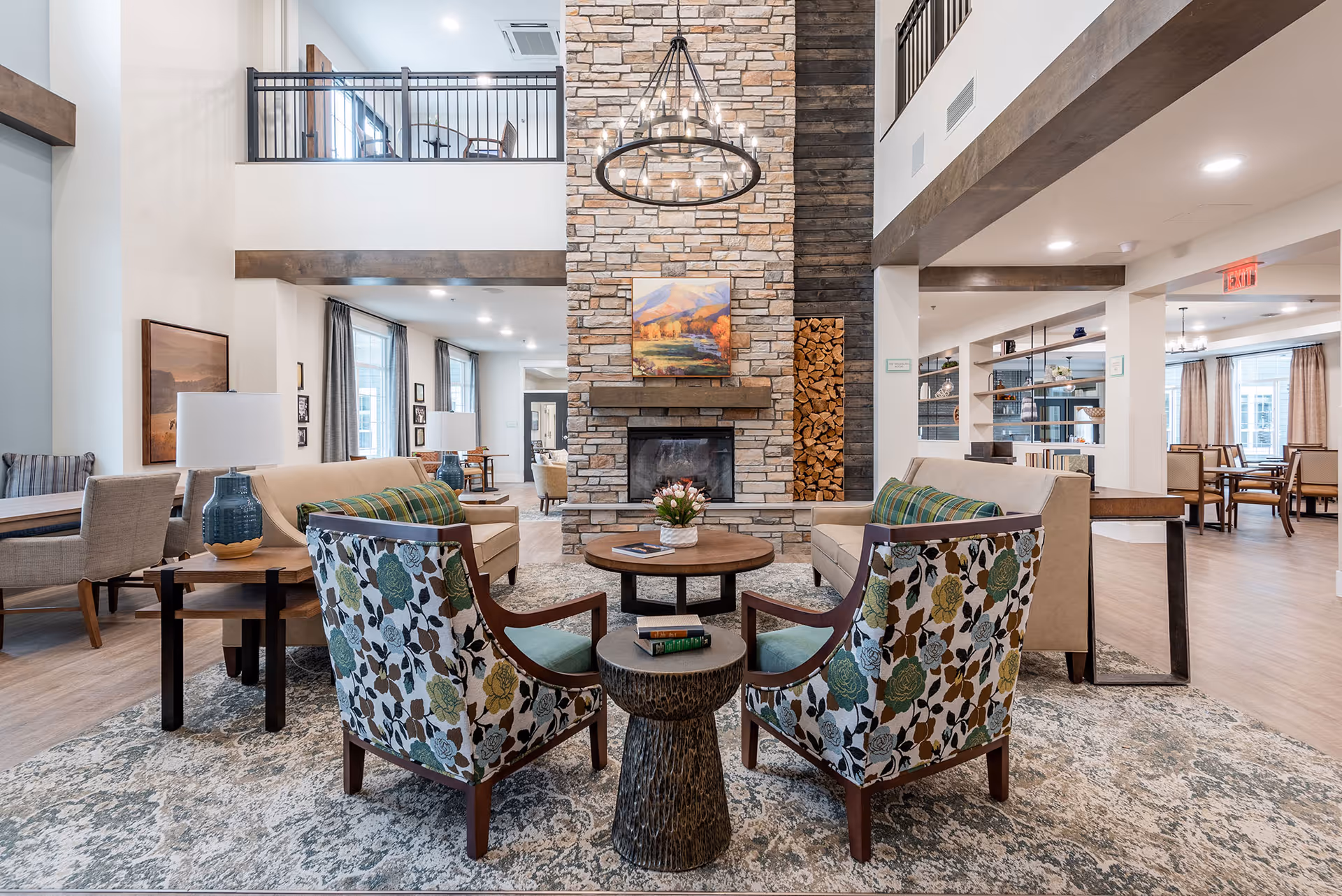 Bright, spacious communal living room with patterned armchairs and sofas arranged around a central stone fireplace beneath a large chandelier.