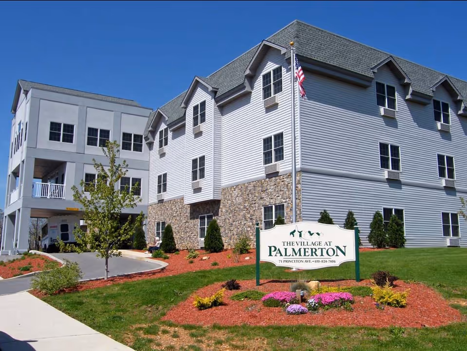 Exterior view of The Village at Palmerton senior living facility showing a multi-story building with white siding and stone accents, a landscaped garden with flowers and shrubs, a flagpole with an American flag, and a sign displaying the facility's name and contact information.