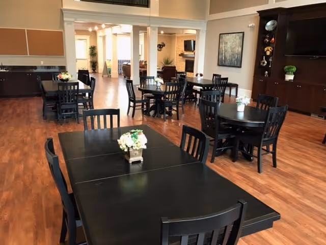 A spacious dining room with multiple black tables and chairs arranged on a wooden floor. Each table has a small floral centerpiece. The room features large windows, a flat-screen TV mounted on a dark wooden cabinet, and light-colored walls with decorative artwork. The area is well-lit with natural light coming from the windows in the background.