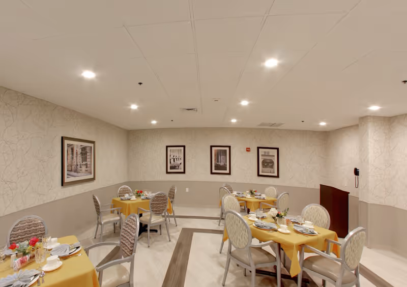 A dining room with several round and rectangular tables covered with yellow tablecloths, each set with plates, cups, and napkins. The room has beige patterned wallpaper, framed pictures on the walls, and recessed ceiling lights. Chairs with patterned upholstery surround the tables.