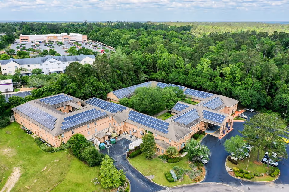 Aerial view of Woodmont Senior Living facility surrounded by trees and greenery, with solar panels installed on the roofs. The building is shaped in a U-formation with a parking lot and driveway in front. In the background, there is a shopping center with a Walmart and other stores.