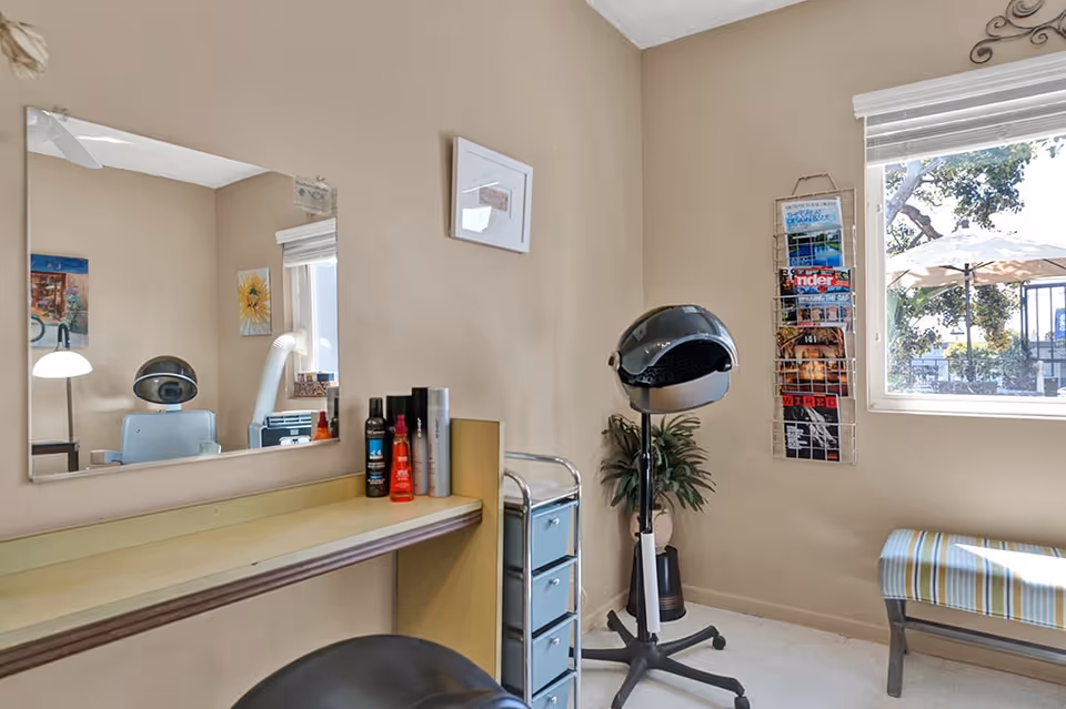 Small salon-style interior with a styling station, mirror, hooded hair dryer, magazine rack and a window with seating.