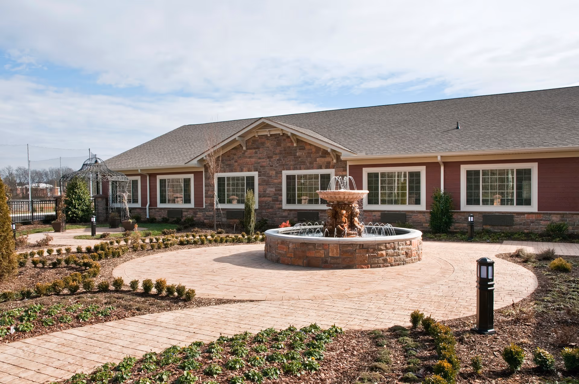 Outdoor courtyard area of a senior living facility with a circular stone fountain in the center, surrounded by landscaped garden beds and a paved walkway. The building in the background has a stone and red siding exterior with multiple windows and a gray shingled roof. There is a metal gazebo structure on the left side and several small outdoor lights along the pathway.