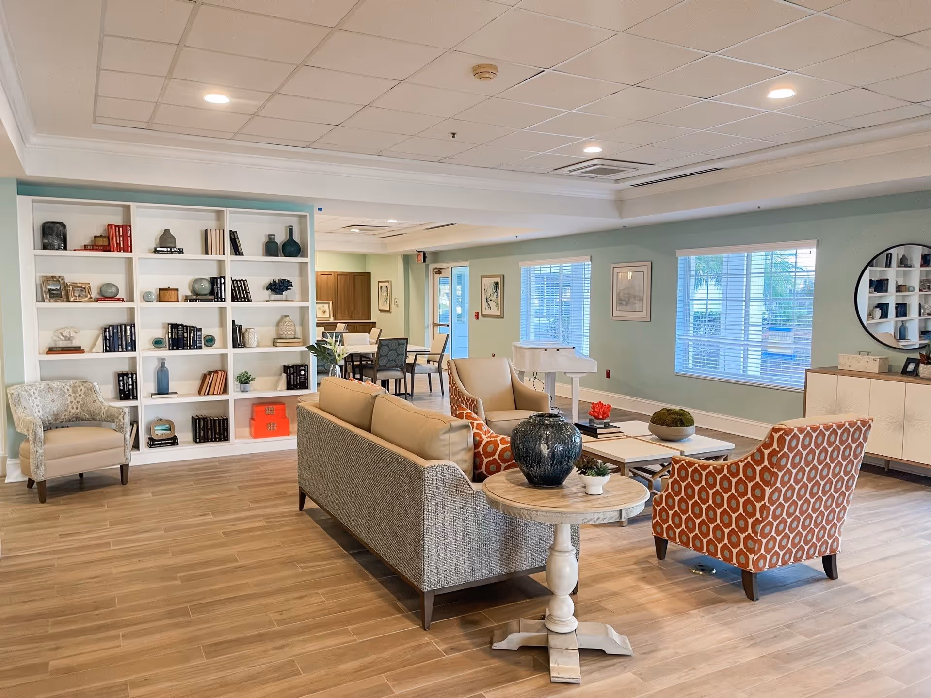 A bright and spacious living room area in a senior living facility with a beige sofa, two patterned armchairs, a round wooden side table with decorative vases, a white coffee table, and a white bookshelf filled with books and decorative items. The room has light wood flooring, light blue walls, large windows with blinds, and a round mirror on the wall. In the background, there is a dining area with tables and chairs.