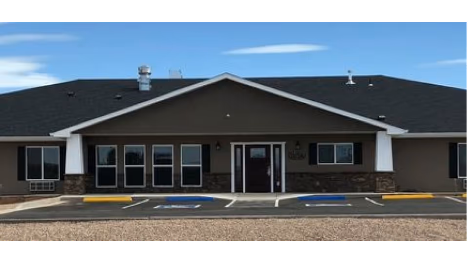 Front exterior view of a single-story building with a dark roof and brown walls, featuring multiple windows and a central entrance door. The parking lot in front has marked handicap parking spaces and yellow curb markings under a blue sky with some clouds.