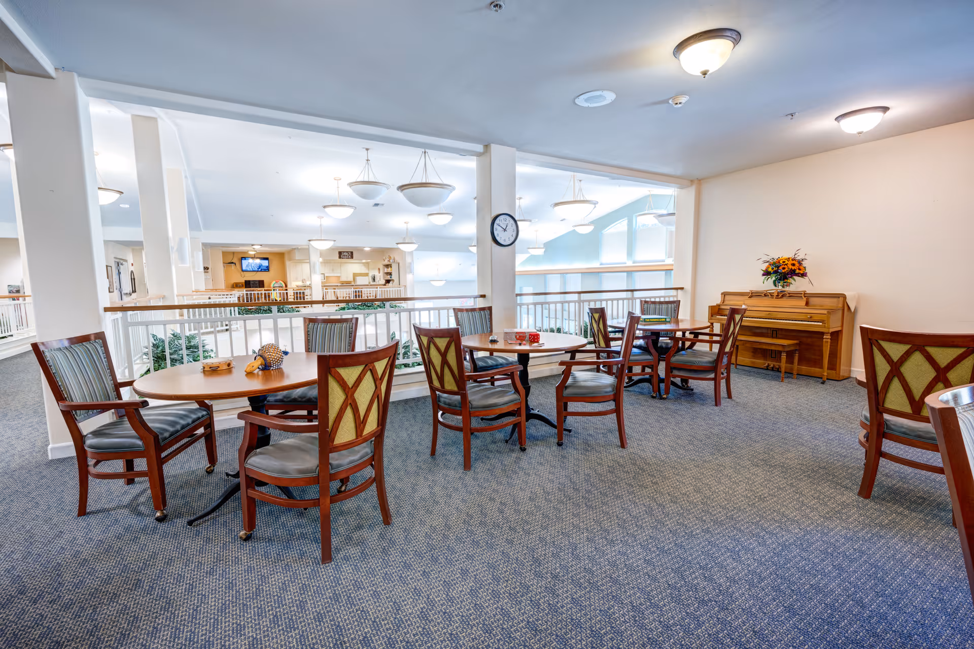 A bright common area in a senior living facility with several round wooden tables and chairs arranged on a carpeted floor. A wooden piano with a flower arrangement on top is against the wall. The area overlooks a lower level with railings and hanging ceiling lights.