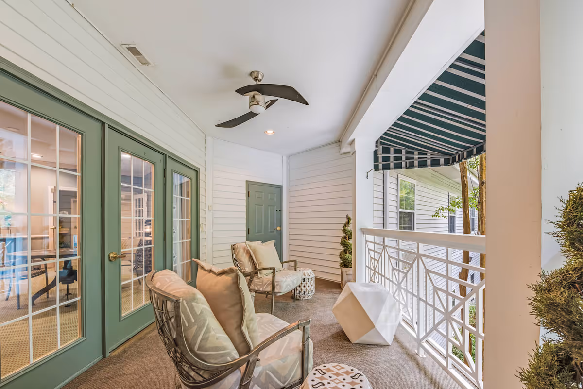 A covered balcony area with two cushioned chairs, a small decorative table, and a geometric white stool. The balcony has white railings and a green and white striped awning. There are green double doors with glass panes leading inside, and a ceiling fan with two blades is mounted on the white ceiling.