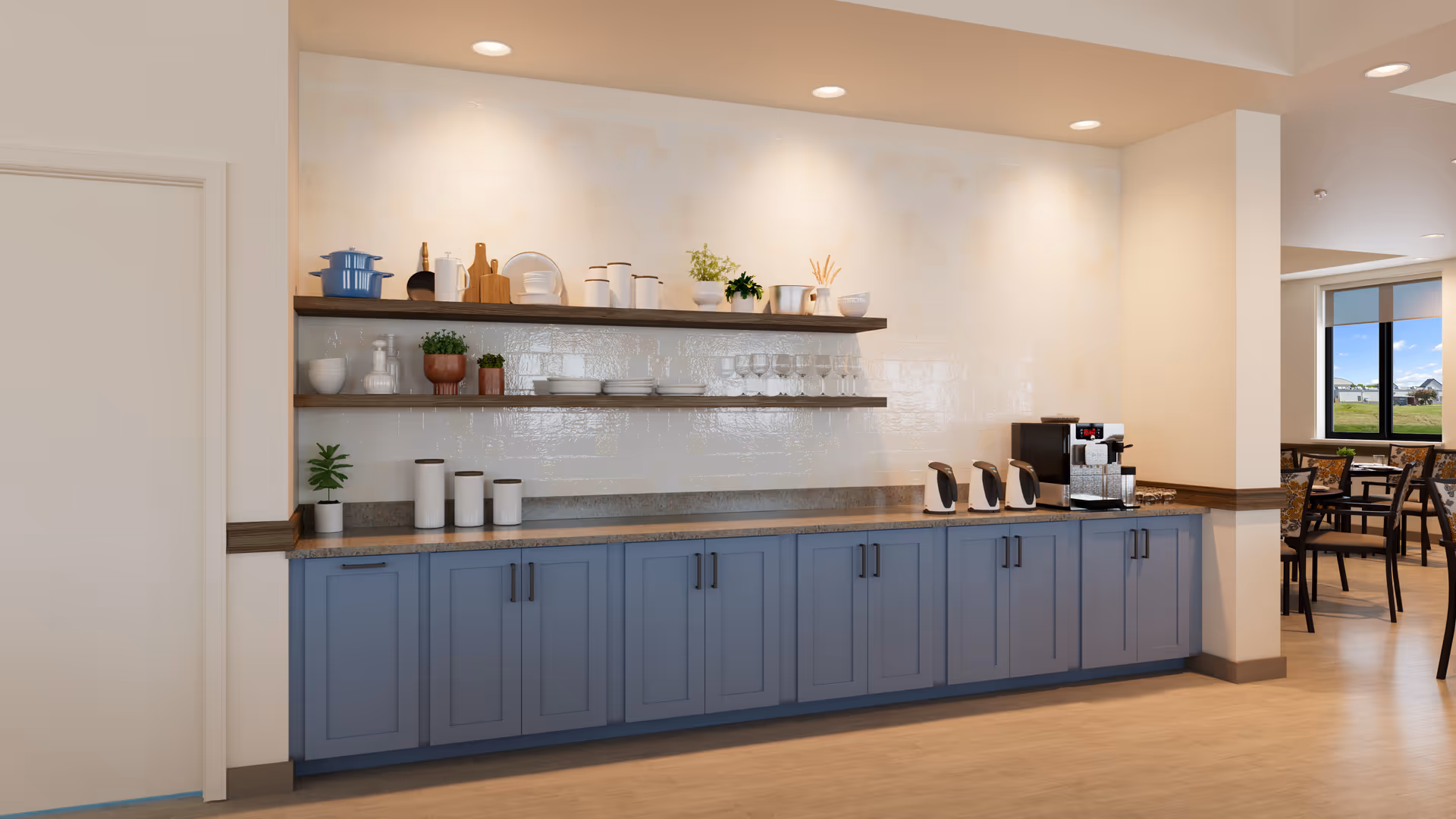 Communal kitchenette with blue lower cabinets, open shelves holding dishes and plants, and a coffee station next to a dining area.
