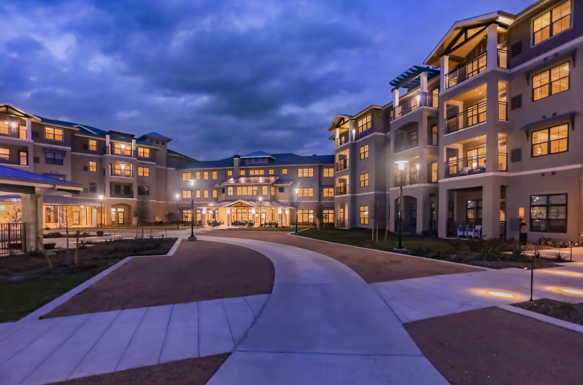 Evening view of a large, multi-story retirement community building with illuminated windows and outdoor lighting along a curved walkway leading to the entrance under a cloudy sky.