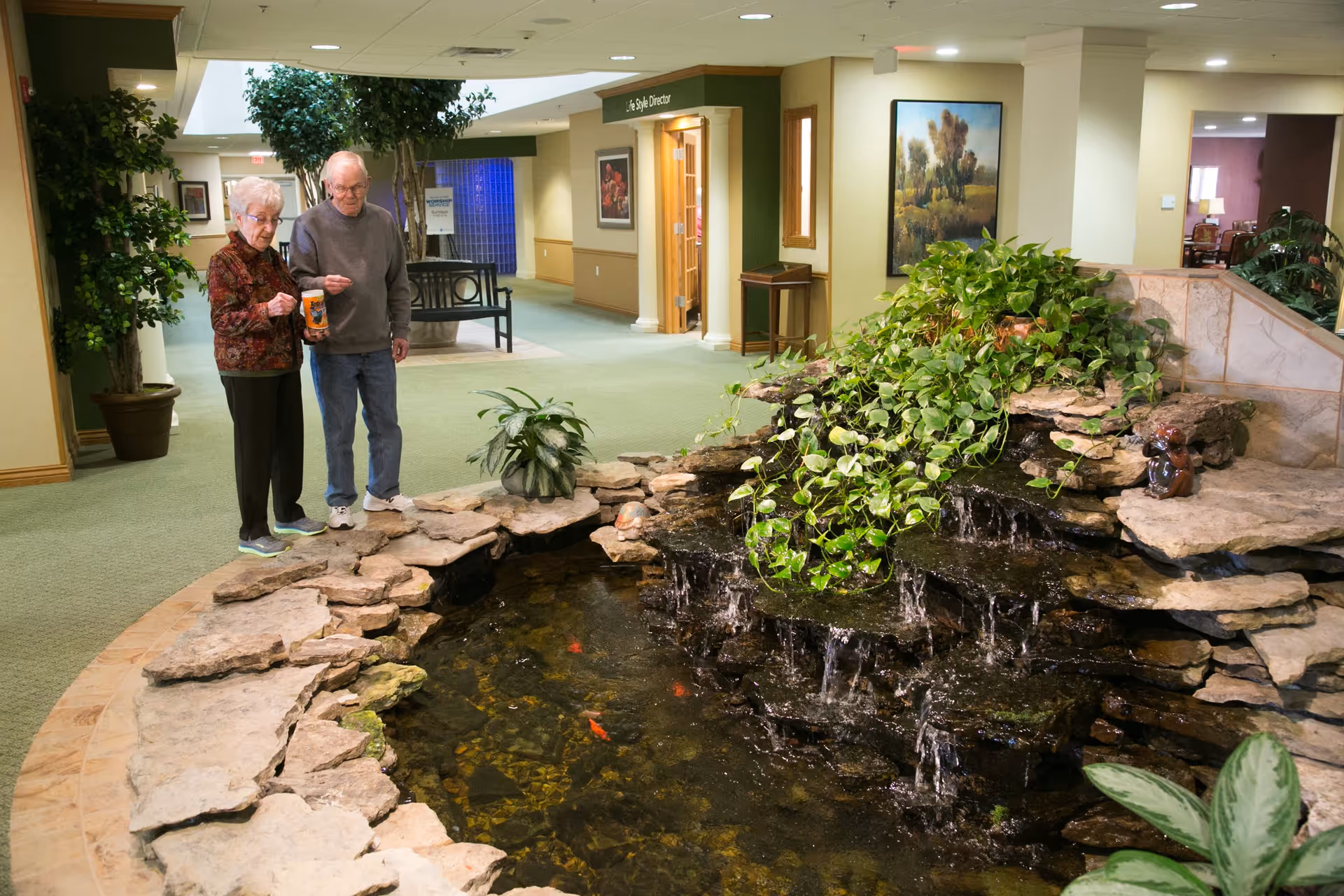 An elderly man and woman standing indoors next to a decorative indoor pond with a small waterfall and green plants. The setting appears to be a common area in a retirement facility with carpeted floors, potted plants, and framed artwork on the walls.