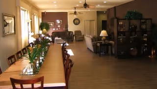Open communal dining and living area featuring a long wooden dining table with chairs and flowers, plus sofas, lamps, and shelving.