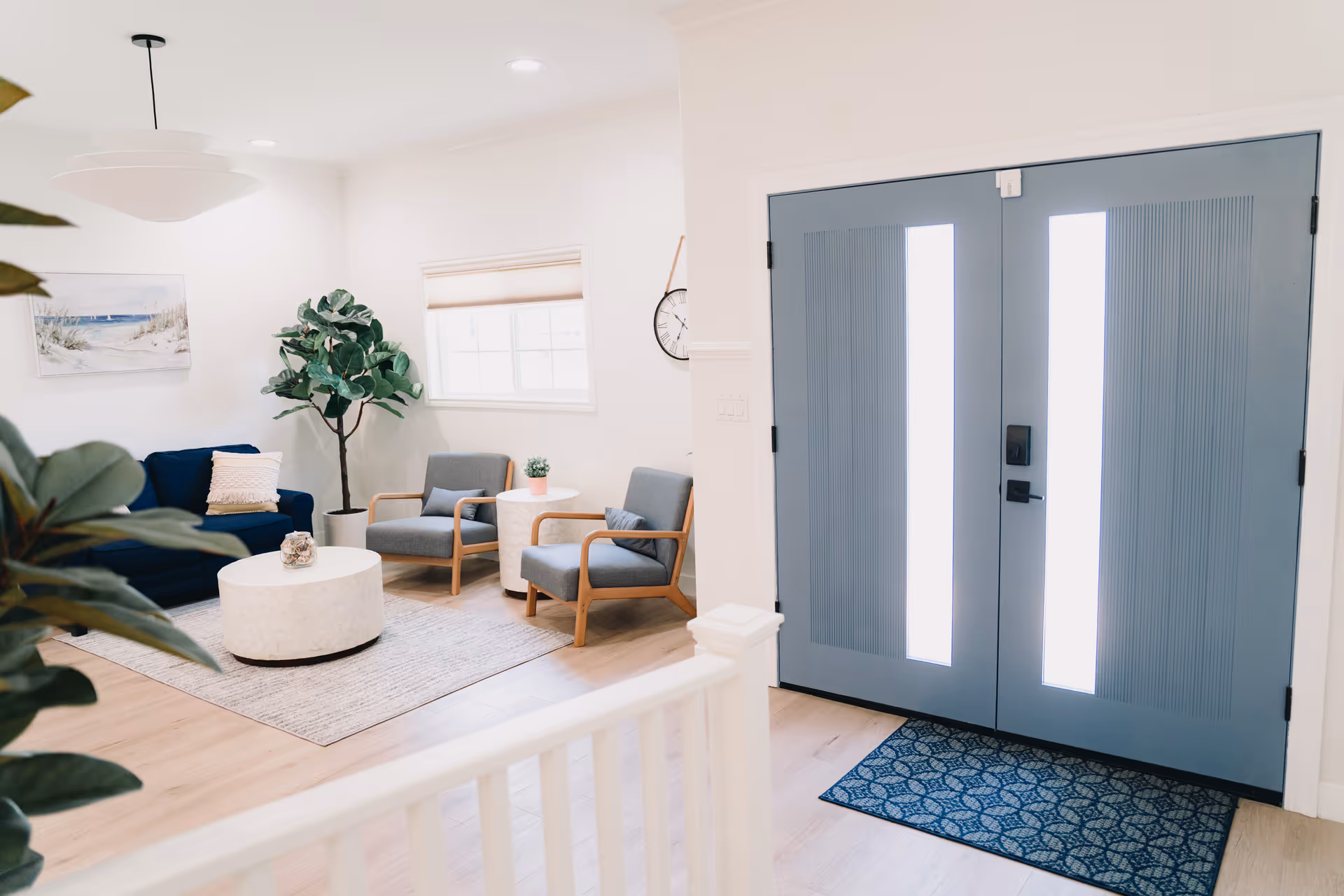 Bright and modern living room area with a blue couch, two gray armchairs with wooden arms, a round white coffee table on a light gray rug, a large potted plant, a wall clock, and a window with a beige blind. To the right, there is a double blue front door with vertical glass panels and a blue patterned doormat.