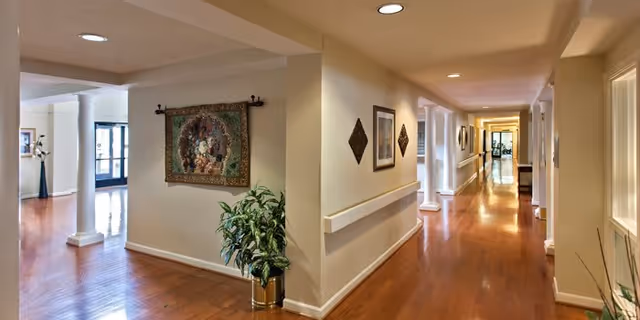 Interior view of a well-lit hallway in a senior living facility with polished wooden floors, white columns, handrails along the walls, decorative wall art, and plants in pots. The hallway extends into the distance with doors and windows along the sides.