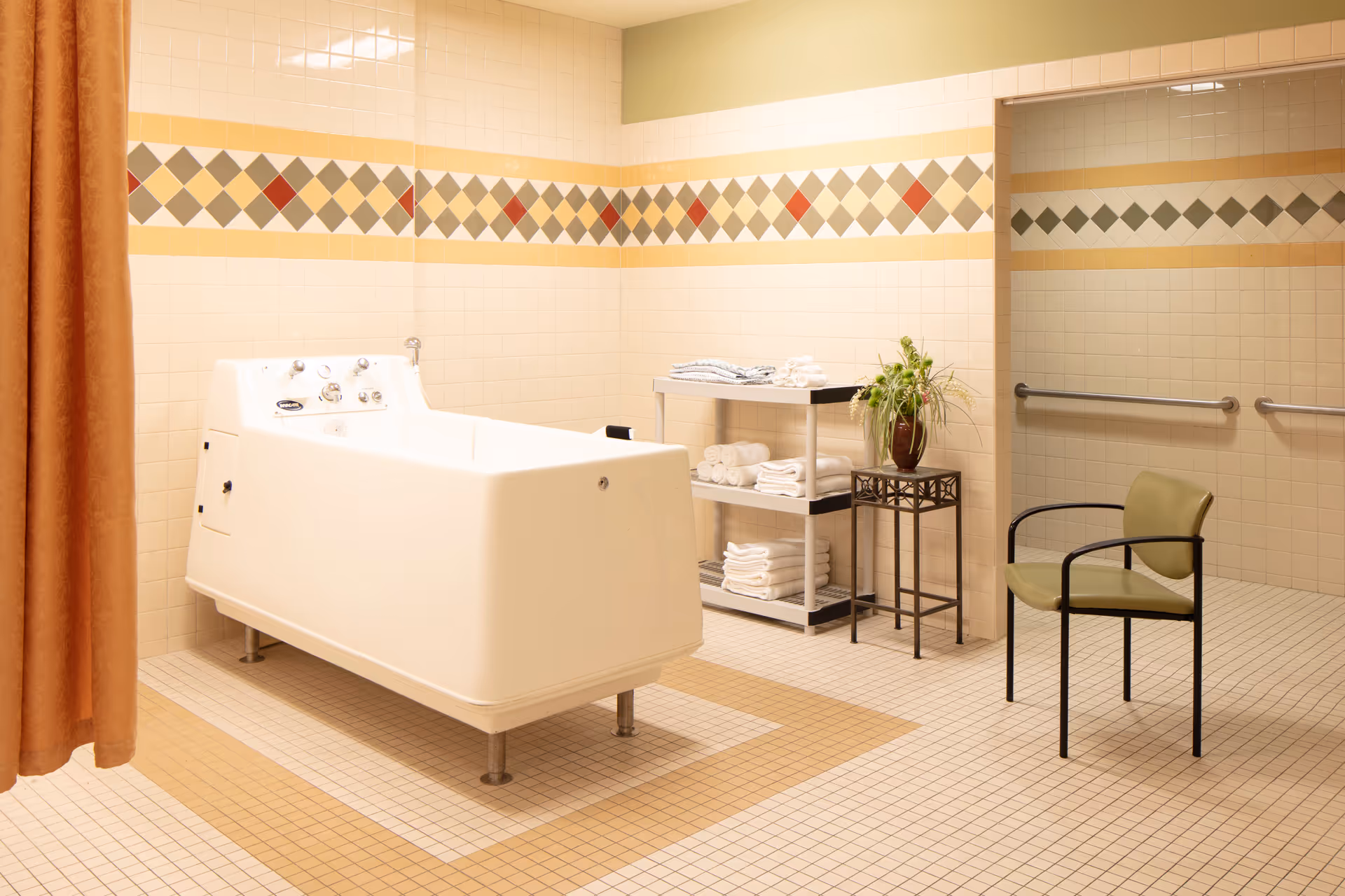 A clean and spacious bathing room with a large white therapeutic bathtub, beige tiled walls and floor with a decorative diamond pattern border, a metal rack holding neatly folded white towels, a small table with a potted plant, a green chair with black metal arms and legs, and an orange privacy curtain partially visible on the left side.