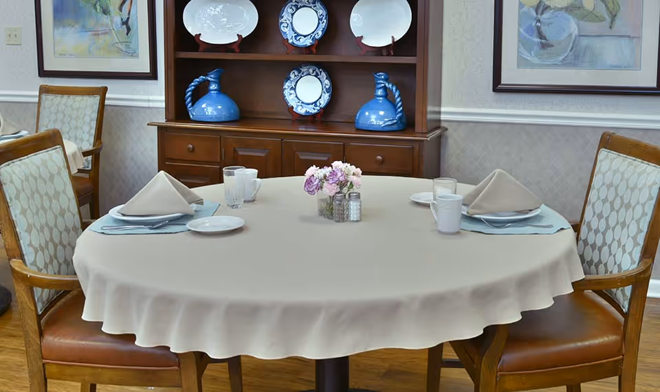 Round dining table set for four with folded napkins, glasses, and a small floral centerpiece in a dining room with a wooden hutch displaying plates.