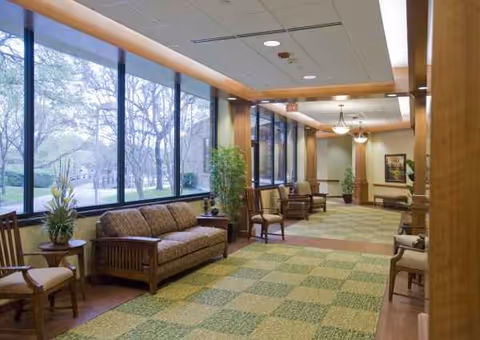 Bright senior living lobby with sofas, chairs, plants, and large windows along the hallway.