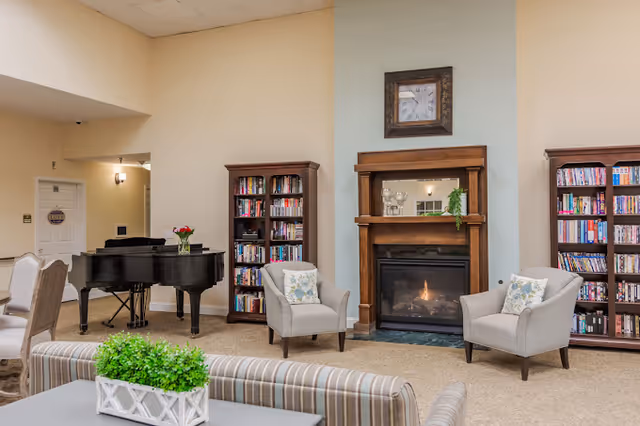 A cozy living room area featuring a lit fireplace with a wooden mantle, flanked by two bookshelves filled with books. Two armchairs with floral cushions are positioned in front of the fireplace. A grand piano is visible in the background near a doorway, and a striped sofa with a green plant on the table in front is partially visible in the foreground.
