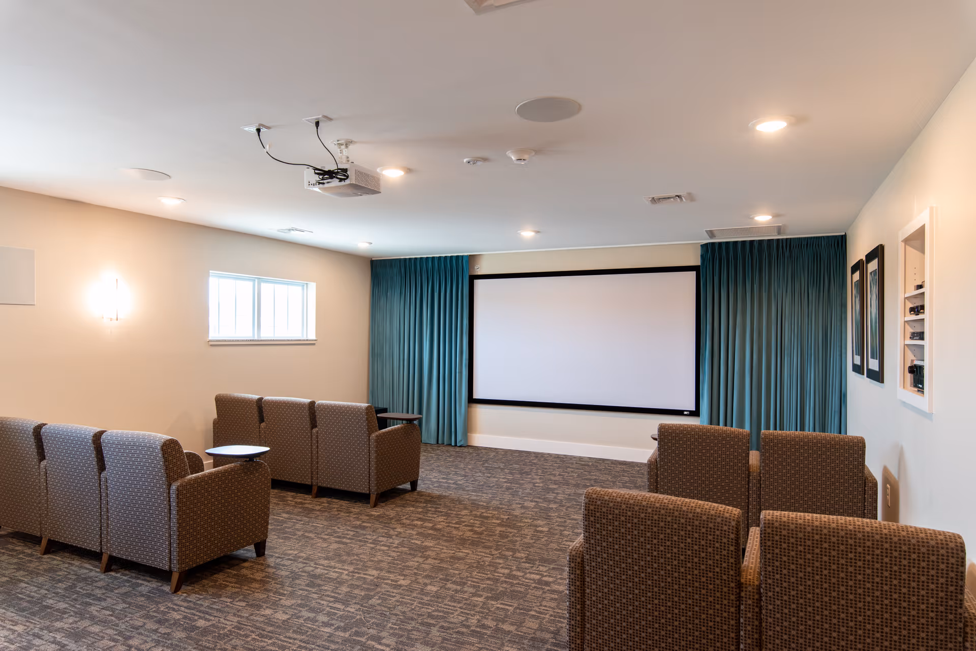 A small theater room with a large white projection screen on the wall, teal curtains on either side, and several rows of brown upholstered chairs facing the screen. The room has beige walls, a carpeted floor, ceiling lights, and a projector mounted on the ceiling.