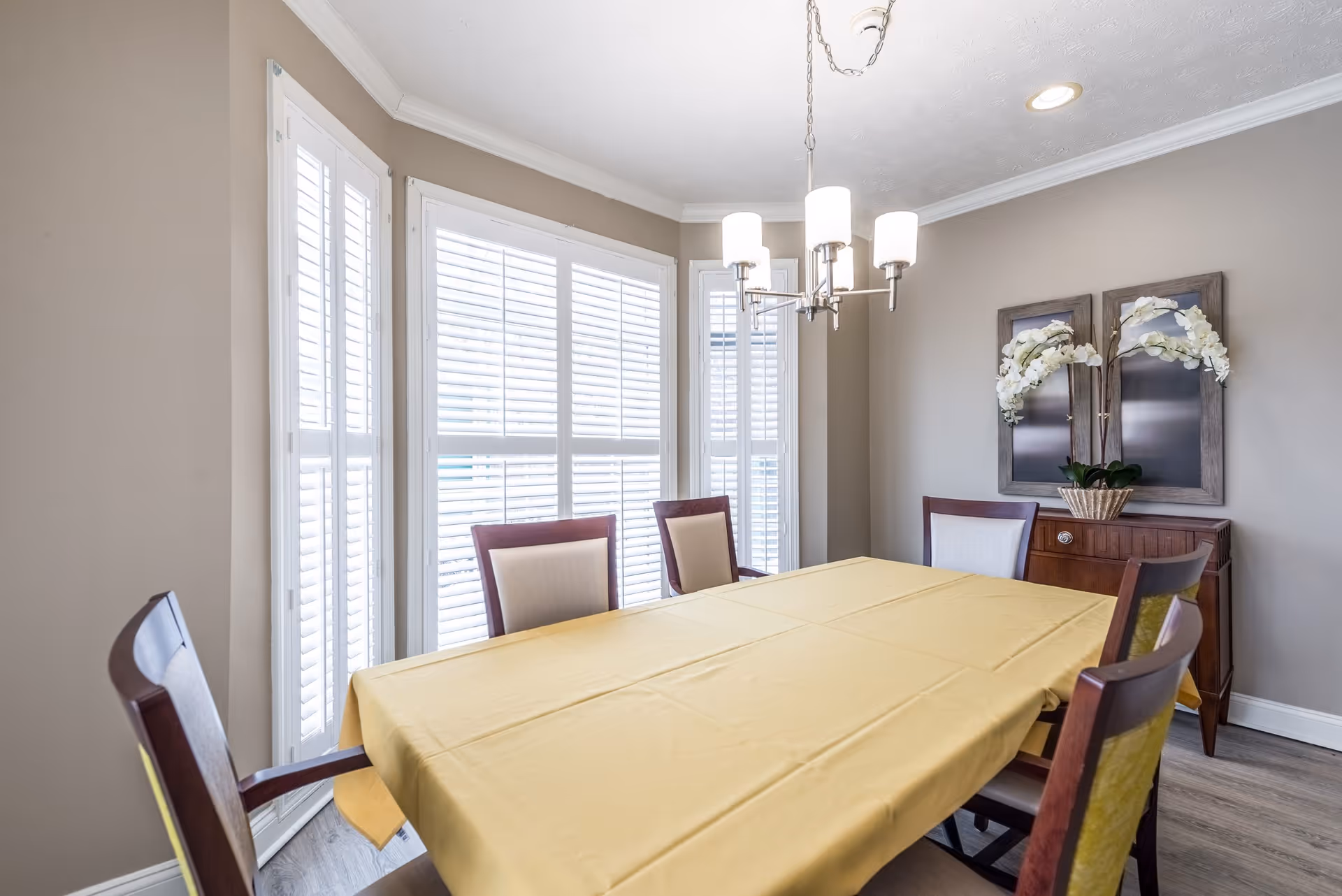 Bright dining room with a yellow-tablecloth-covered table, chairs, bay windows with shutters, a chandelier, and a sideboard with floral decor.