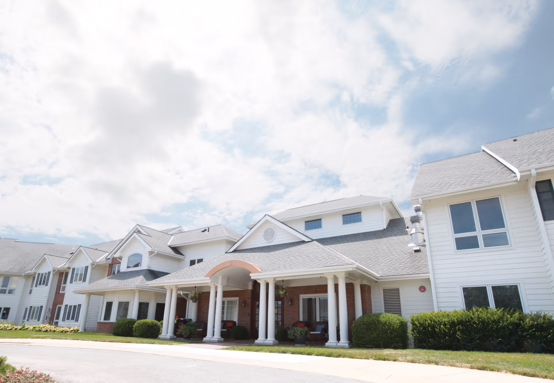 Exterior view of a large assisted living facility building with white siding, multiple windows, and a covered entrance supported by white columns. The sky is partly cloudy and there is a well-maintained lawn and shrubs around the building.