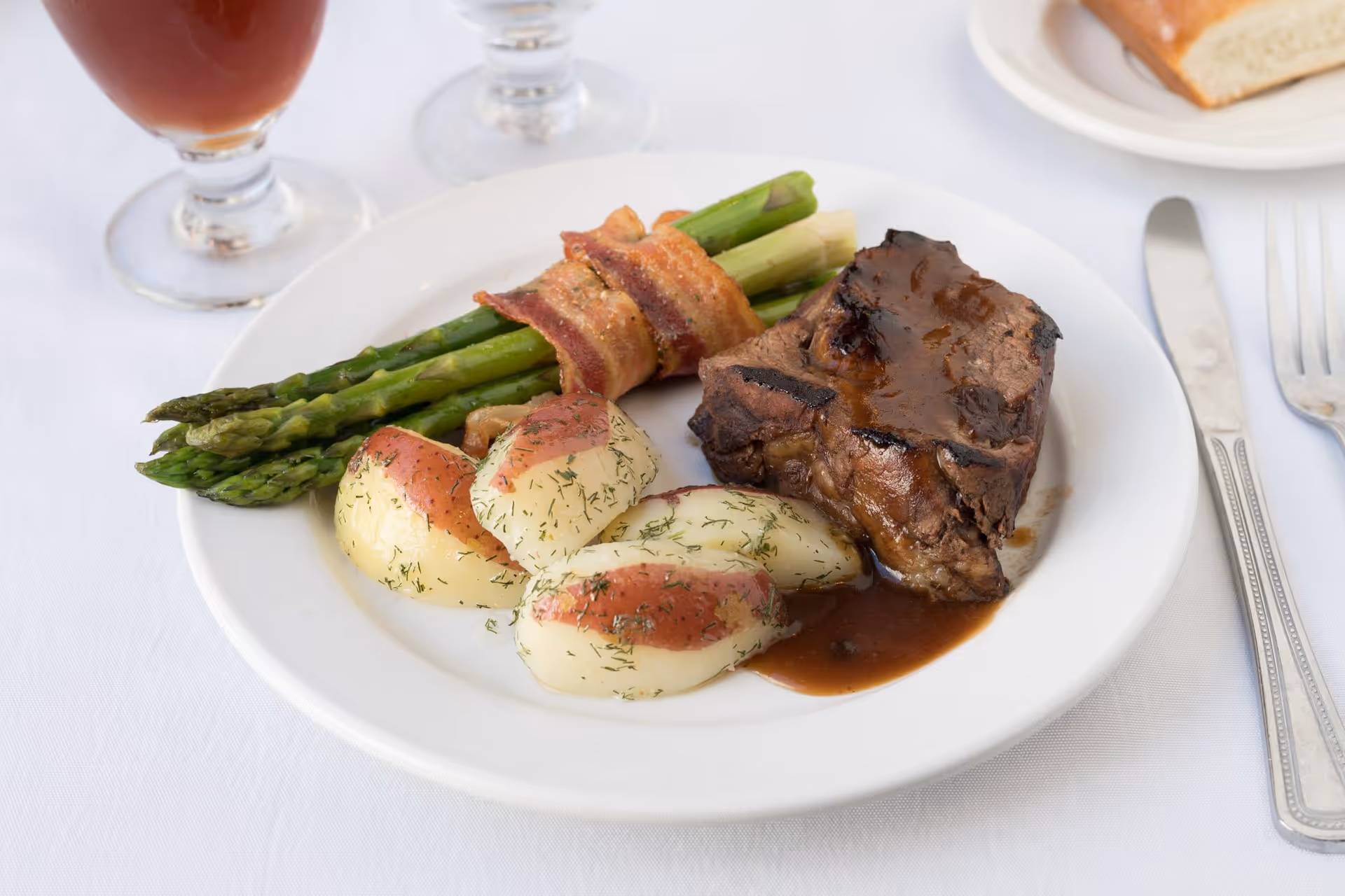 A white plate with a serving of grilled meat covered in brown sauce, three boiled red potatoes sprinkled with herbs, and a bundle of asparagus wrapped in bacon. The plate is on a white tablecloth with a knife and fork to the right, a piece of bread on a small plate in the background, and two glasses partially visible.