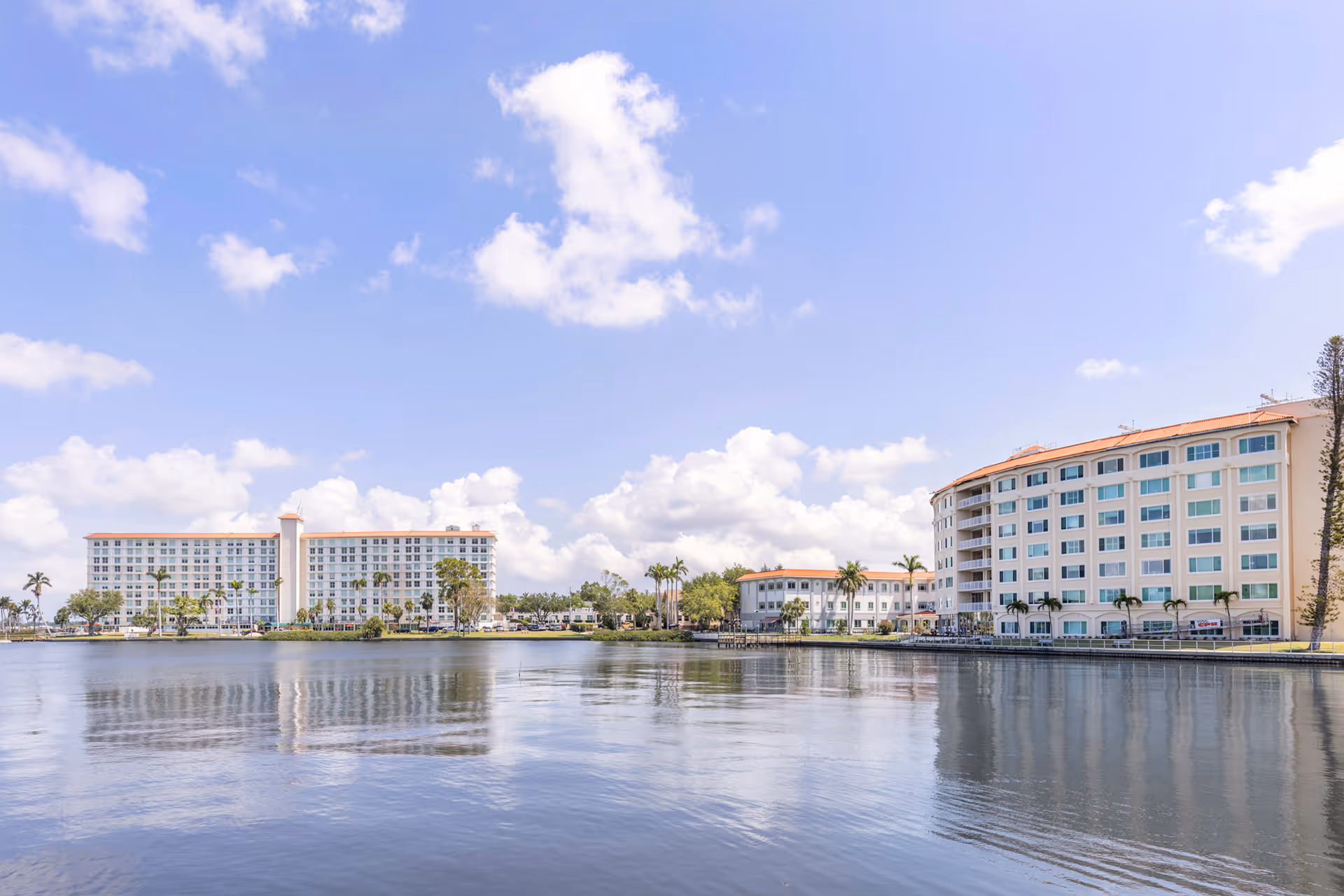 View of two large multi-story buildings by a calm body of water under a partly cloudy blue sky, with palm trees and greenery around the buildings.