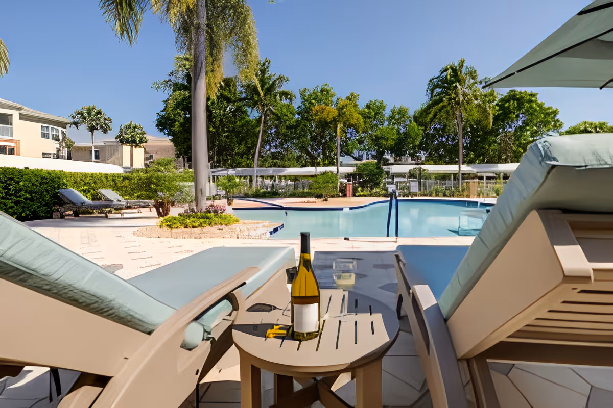 View of an outdoor pool area with two lounge chairs and a small table between them holding a bottle of wine and a glass. The pool is surrounded by trees, bushes, and additional lounge chairs under a clear blue sky.