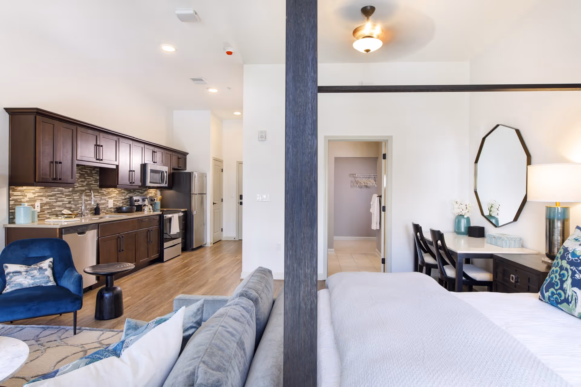 Interior view of a modern assisted living apartment showing a kitchen with dark wood cabinets, stainless steel appliances, and a living area with a blue armchair and gray sofa. Adjacent to the living area is a bedroom with a bed, a desk with two chairs, a lamp, and a mirror on the wall. The space has light-colored walls and wood flooring.