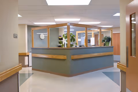 Interior view of a reception or nurse station area in a senior living facility with a central desk enclosed by glass panels. The area has beige walls, a clock on the wall, and plants on the desk. The floor is tiled with a pattern of white and green sections, and wooden handrails are mounted on the walls.