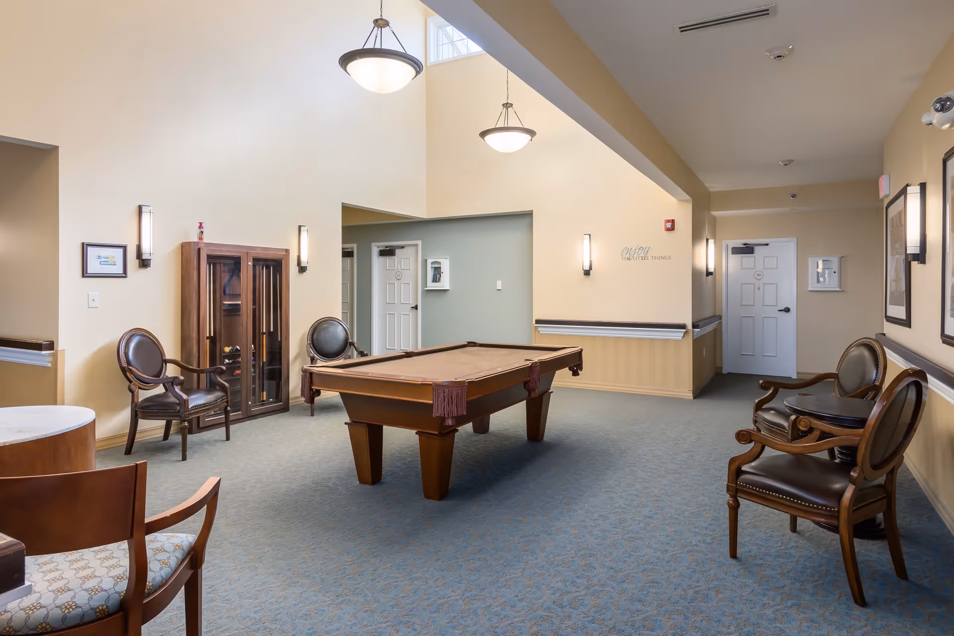 A spacious room with a pool table in the center, surrounded by several wooden chairs with leather cushions. The room has beige walls, blue carpet flooring, and two hanging ceiling lights. There is a wooden cabinet against the wall and framed pictures on the walls. A door is visible at the far end of the room.