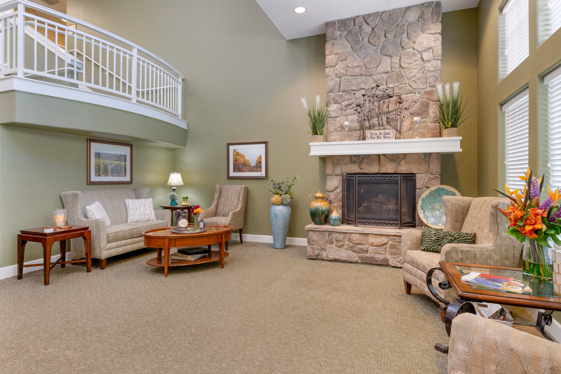 A cozy living room area with a stone fireplace, beige carpet, and green walls. The room features a beige sofa, an armchair, a wooden coffee table, side tables with lamps, decorative vases, framed artwork on the walls, and a large window with white blinds letting in natural light. There is a white railing balcony above the sofa area.