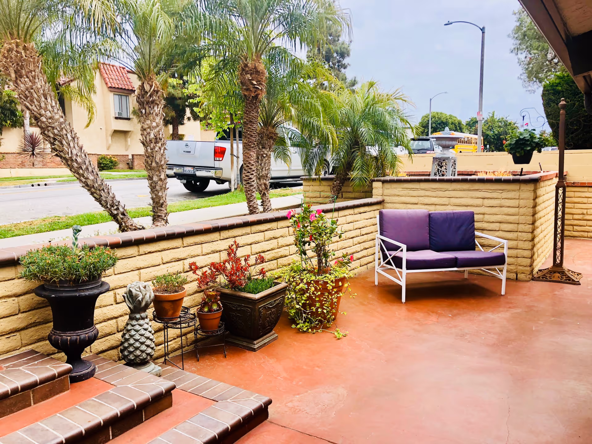 Outdoor patio area with terracotta flooring, a small purple cushioned bench, several potted plants including palm trees and flowering plants, a decorative pineapple statue, and a beige brick wall surrounding the space. A white pickup truck and a yellow school bus are visible on the street beyond the patio.