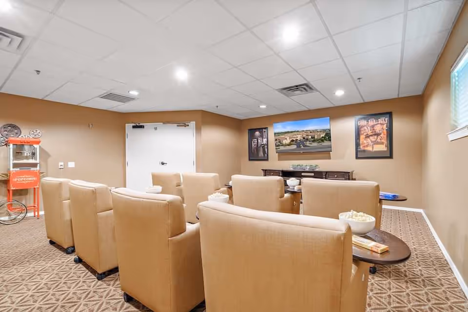 Small media room with rows of tan armchairs facing a wall-mounted TV and a popcorn machine in the corner.
